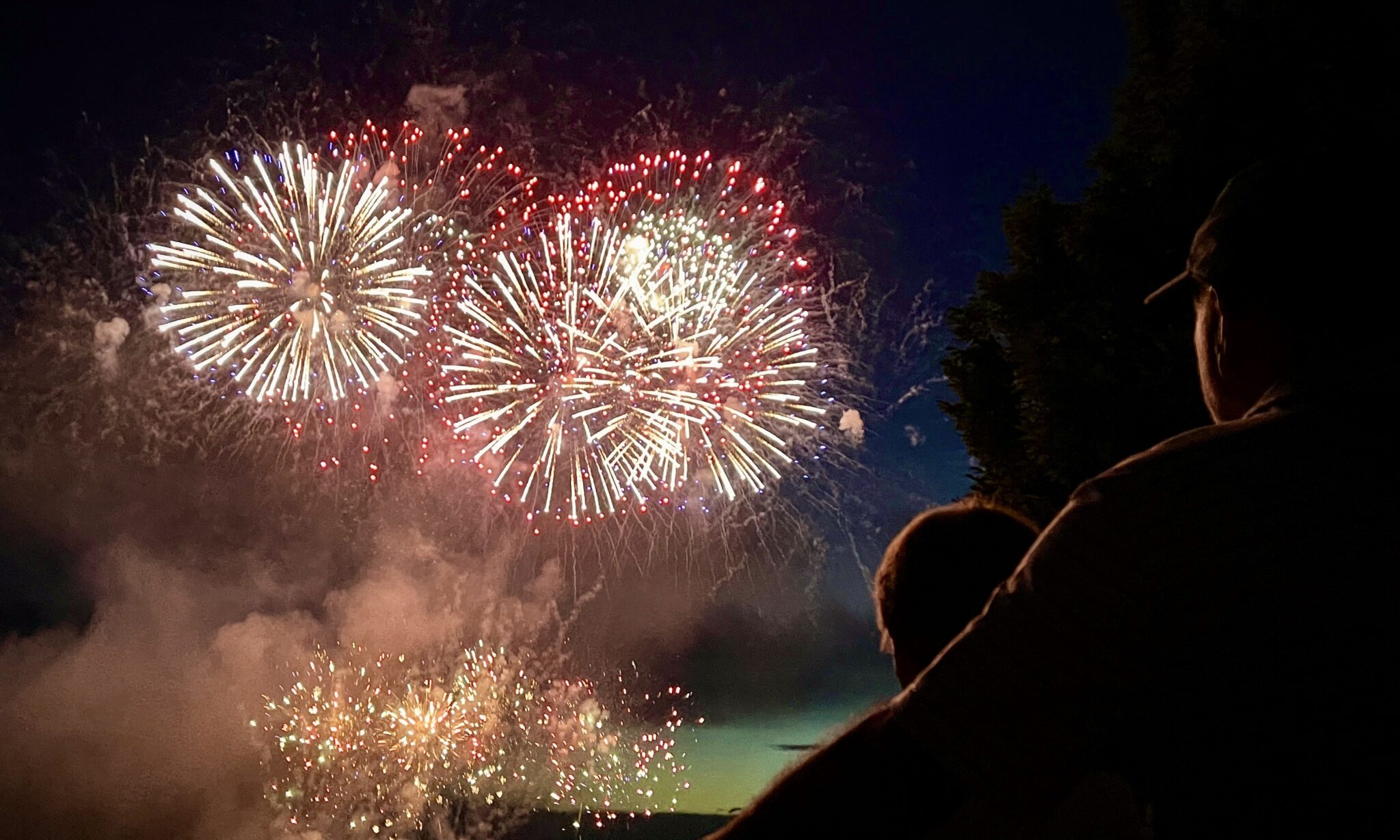 Fireworks explode in the sky while a man has his arm behind a shorter woman while both looking on. The light from the fireworks illuminate the sides of their heads and faces. 