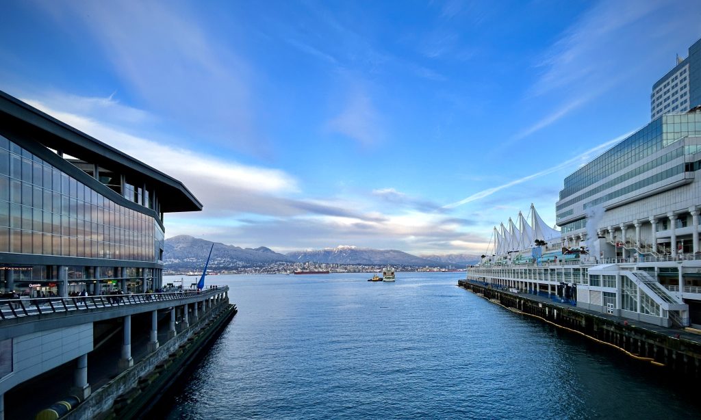 A landscape photo from downtown Vancouver, B.C. that looks across the Burrard Inlet and taken between Canada Place on the right and the Vancouver Convention Centre on the left. The sky is blue with wispy clouds and the mountains on the North Shore are powdered with snow in the distance.
